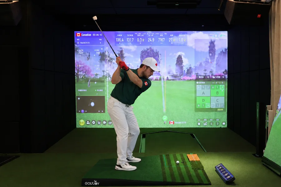 Golfer at the top of his backswing in a simulator bay with impact screen, hitting mat, auto tee-up and remote